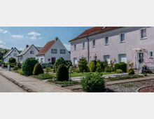 Straßenansicht einer Wohnsiedlung mit Reihenhäusern und Einfamilienhäusern, umgeben von gepflegten Vorgärten mit Sträuchern und Rasenflächen; gepflasterte Gehwege verlaufen entlang der Straße.