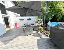 Holzterrasse an einem Wohnhaus mit großem Ampelschirm, Rattan-Sitzgruppe und Pflanzkübeln vor einer weißen Mauer mit Blumenkästen. Rechts stehen ein aufstellbarer Pool neben dichtem Grün sowie ein abgedeckter Grill.