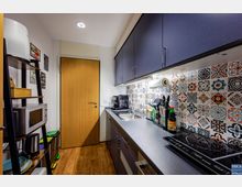 Kleine, moderne Küche mit einem Holzfußboden, dunklen Schränken und einer Arbeitsplatte. Die Rückwand ist mit bunten, gemusterten Fliesen verkleidet; an der rechten Seite befinden sich ein Herd, eine Spüle und Küchengeräte, links ein Regal mit Mikrowelle und Ablagefächern.