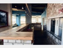 Industrieähnlicher Innenraum mit einer Holztheke auf gemauertem Unterbau, einer kleinen Spüle und Hängelampen an der Decke. Rechts befinden sich ein Fahrstuhlschacht sowie unverputzte Wände aus Beton und Ziegeln; helle Fensterflächen im Hintergrund lassen Tageslicht herein.