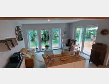 Wohnzimmer mit großen bodentiefen Fenstern und Glastüren, die einen Blick auf einen Garten mit Bäumen und eine Terrasse ermöglichen. Der Raum ist mit einem Sofa, Sesseln, einem Teppich und Zimmerpflanzen eingerichtet, während dekorative Elemente wie Bilderrahmen und Regale die Wände und Möbel schmücken.