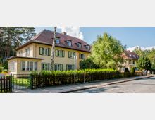 Straßenansicht eines Wohnviertels mit zweigeschossigen gelben Häusern und roten Ziegeldächern, umgeben von gepflegten Vorgärten mit Hecken und einem Holzzaun. Im Hintergrund sind Bäume und ein blauer Himmel mit wenigen Wolken sichtbar.