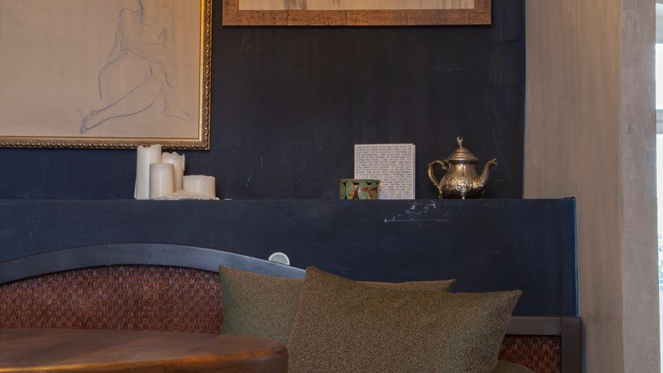 Gemütliche Sitzecke in einem Innenraum mit dunkler gepolsterter Bank, Kissen und rundem Holztisch. An der Wand hängen gerahmte Zeichnungen, darüber hängt eine große kugelförmige Hängelampe; auf einer Ablage stehen Kerzen und eine Teekanne.