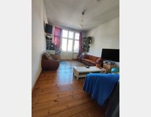 Long, narrow living room with wooden plank flooring, white walls, and a central ceiling light. Large multi-pane windows and a glass door at the far end are framed by red curtains, with sofas along the walls, a TV on a stand, and a white coffee table in the middle.