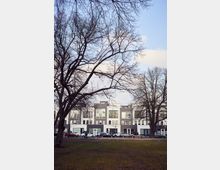 A row of modern white and gray apartment buildings with large windows, viewed from across a park with bare trees and a grassy area in the foreground. Parked cars line the street in front of the buildings, and the sky above is partly cloudy.