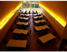 Innenraum einer kantinenähnlichen Einrichtung mit mehreren rechteckigen Tischen und schwarzen Stühlen, die in Reihen angeordnet sind. Das hohe, schrägdachartige Zimmer ist mit gelber Beleuchtung und einer großen Fensterfront im Hintergrund ausgestattet, die einen Blick ins Grüne bietet.