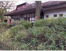 Fachwerkhaus mit einem roten Ziegeldach und Dachgauben, umgeben von dichter Vegetation und einem niedrigen Holzzaun. Ein großer Baum steht vor dem Gebäude, und im Hintergrund sind Terrassenmöbel sowie Sonnenschirme sichtbar.