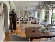 Wohnzimmer in einem gehobenen Stil mit Holzboden und einem großen Teppich. Der Raum enthält eine gemütliche Sitzecke, Wanddekoration mit gerahmten Bildern, eine gläserne Doppeltür zum Balkon und einen Holztisch mit Stühlen im Vordergrund.