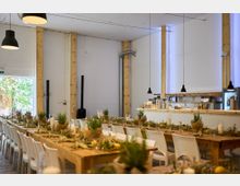 Ein heller, moderner Veranstaltungsraum mit einer einfachen Holzkonstruktion, langen Holztischen und weißen Stühlen. Die Tische sind dekoriert mit grünen Pflanzen, Kerzen und Naturmaterialien, während im Hintergrund eine minimalistisch gestaltete Bar zu sehen ist.