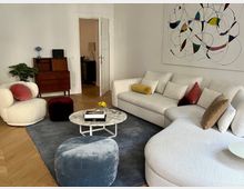 Wohnzimmer mit einer weißen, geschwungenen Couch, einem Marmortisch und einem weißen Sessel mit rosa Kissen. Im Hintergrund stehen eine Holzkommode, eine grüne Zimmerpflanze und eine Tür, die in einen weiteren Raum führt. An der Wand hängt ein großes abstraktes Gemälde mit bunten Formen und Linien.