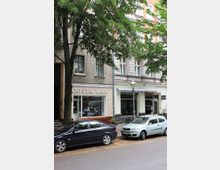 Straßenszene mit einer Baumreihe und geparkten Autos vor einer Reihe von Wohn- und Geschäftshäusern. Im Vordergrund ist ein Schaufenster mit der Aufschrift „CRYSTAL BALL“ zu sehen, daneben ein Geschäft mit gestreifter Markise und ein Schild mit der Aufschrift „BAR“.