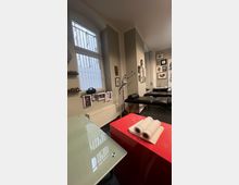 Raum eines Tattoostudios mit cremefarbenen Wänden, großen vergitterten Fenstern und einer roten Arbeitsfläche im Vordergrund mit Papiertüchern. Im Hintergrund sind eine Behandlungsliege, Kunstwerke an den Wänden und Lampen zur Beleuchtung sichtbar.