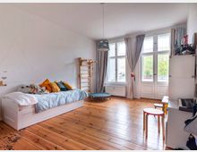 Ein schlicht eingerichtetes Kinderzimmer mit Holzdielenboden, einem weißen Einzelbett mit Kuscheltieren, einem Klettergerüst aus Holz an der Wand und einer Sitzecke mit Holzhockern. Große Fenster und Glastüren mit langen Vorhängen lassen viel Tageslicht herein und bieten Blick auf draußen.