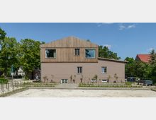 Zweistöckiges Gebäude mit modernem Design; die untere Etage ist beige verputzt, die obere Etage hat eine Holzfassade. Im Vordergrund ein gepflasterter Hof mit jungen Pflanzen, im Hintergrund Bäume und Nachbarhäuser.