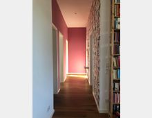 Ein Flur in einem Wohnbereich mit rosa gestrichenen Wänden, einem Bereich mit floraler Tapete und Holzboden. Rechts ist ein Bücherregal sichtbar, links führen mehrere Türen in angrenzende Räume.