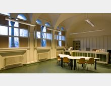 Innenansicht eines Verwaltungs- oder Arbeitsraums mit hohen, gewölbten Decken. Große Fenster mit Bogenform, ein zentral stehender Tisch mit Stühlen und ein Empfangsbereich mit Schreibtischen und Bürostühlen prägen die Gestaltung.