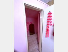 Blick in einen kleinen, rosa gestrichenen Raum mit Holzboden und einem runden Türbogen im Hintergrund. An der weißen Tür im Vordergrund hängt eine Kette aus pinkfarbenen Pompons.