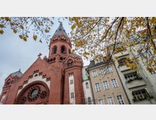 Backstein-Kirche im neugotischen Stil mit großem Turm und Rundfenstern, daneben stehen mehrstöckige Wohnhäuser mit Balkonen. Herbstbäume mit gelben Blättern ragen in das Bild.