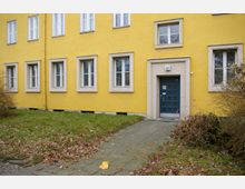 Gelbes Wohngebäude mit weiß gerahmten Fenstern und einer dunkelblauen Eingangstür. Vor dem Gebäude führt ein gepflasterter Weg durch eine mit Laub bedeckte Grünfläche.