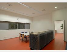 Ein moderner Raum mit rotem Boden, einem rechteckigen Tisch und mehreren Stühlen. Die Wände sind weiß, es gibt eine große Fensterfront mit Rollos, eine Wanduhr und eine offene Tür, die in einen angrenzenden Raum mit einer Pflanze führt.