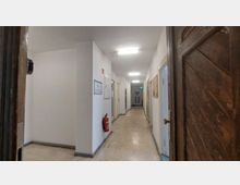 Langer, weißer Gebäudeflur mit Türen zu mehreren Räumen auf beiden Seiten, einer Feuerschutztür am Ende und einem Feuerlöscher an der Wand. Die Decke ist mit rechteckigen Leuchtstofflampen ausgestattet, der Boden besteht aus hellgrauem Material.