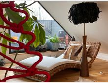 Gemütlicher Wohnraum unter einer Dachschräge mit einem großen, dreieckigen Fenster. Im Vordergrund eine geflochtene Liege mit weißem Kissen und Fellkissen sowie einer Stehlampe mit schwarzem Feder-Lampenschirm, umgeben von Pflanzen und Holzboden.