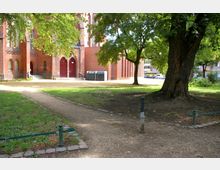 Grünfläche mit Kieswegen vor einer roten Backsteinkirche mit gotischen Elementen, darunter spitzbogige Fenster und Türen. Im Vordergrund stehen große Bäume, die Schatten auf den Platz werfen.