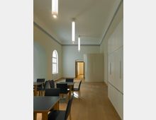 Ein heller Innenraum mit Holzfußboden, Tischen und Stühlen, einer gewölbten Wand mit eingebauten Schränken und großen Fenstern, die natürliches Licht hereinlassen. Moderne Hängelampen beleuchten den Raum, der durch eine geöffnete Tür zu einem weiteren Bereich führt.