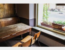 Ein schlichter Innenraum mit einem alten Holztisch, zwei Holzstühlen mit Metallrahmen und einem großen Fenster, das auf die Straße blickt. Auf der Fensterbank stehen Blumenkästen mit Pflanzen, dahinter sind unscharf Fahrräder und Gebäude erkennbar.