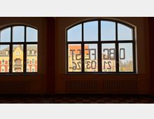 Blick aus einem Innenraum mit zwei großen, segmentbogigen Sprossenfenstern und Heizkörpern darunter. Durch die Fenster sieht man gegenüberliegende Wohnhäuser mit farbigen Fassaden und Satteldächern; auf dem rechten Fenster sind spiegelverkehrte Ziffern und Buchstaben aufgeklebt.
