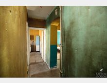 Schmaler, verwitterter Flur in einem Gebäude mit farblich abblätternden Wänden in Grün und Gelb, offenem Zugang zu angrenzenden Räumen und sichtbaren Rohrleitungen an der Wand. Im Hintergrund sind ein heller Raum und ein Kühlschrank erkennbar.