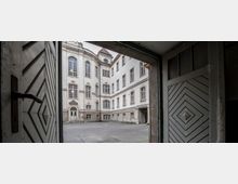 Blick durch ein geöffnetes Holztor auf einen historischen Innenhof mit einem mehrstöckigen Gebäude aus heller Fassade, großen Fenstern und leicht barockem Stil. Der gepflasterte Hof ist leer, und die Architektur wirkt symmetrisch mit rundlichen Gebäudeecken.