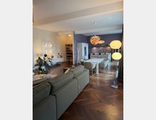 Offener Wohnraum mit modernem Interieur: Im Vordergrund eine graue Polstercouch auf einem Parkettboden mit Fischgrätenmuster, im Hintergrund ein Essbereich mit einem weißen Esstisch und gepolsterten Stühlen. Eine offene Küche mit dunkler Rückwand und warm beleuchteten Hängelampen grenzt an den Essbereich.