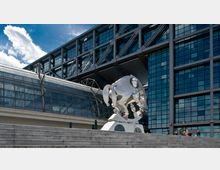 Eine silberne Skulptur in abstrakter Form steht vor dem modernen Hauptbahnhof mit Glasfassade und Stahlkonstruktionen. Im Vordergrund sind Stein-Treppenstufen zu sehen, während der Himmel teils bewölkt ist.