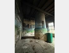 Innenraum eines alten Industriegebäudes mit rußgeschwärzten Ziegelwänden und freiliegenden Deckenbalken. In der Mitte steht ein großer zylindrischer Schornstein- oder Ofenschacht aus Ziegeln, daneben ein grüner Kunststoffbehälter am Fenster.