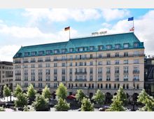Fassade des Hotel Adlon in Berlin, ein mehrstöckiges Gebäude mit cremefarbener Außenwand, zahlreichen Fenstern und einem markanten grünen Mansardendach. Vor dem Gebäude befinden sich Bäume, Gehwege und geparkte Autos; zwei Flaggen, darunter die deutsche, wehen auf dem Dach.