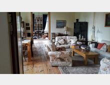 Wohnzimmer in einem klassischen Stil mit Holzfußboden und Teppich. Der Raum enthält floral gemusterte Sofas, einen Holztisch mit Büchern, eine dunkle Vitrine und Lampen; im Hintergrund ist ein angrenzender Raum mit Büchern und einem Schreibtisch sichtbar.