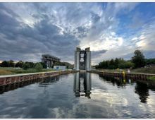 Ein moderner Schiffshebewerk mit Beton- und Stahlstrukturen an einem ruhigen Wasserkanal, eingerahmt von Bäumen und Vegetation unter einem bewölkten Himmel. Links im Bild steht ein technisches Gebäude, während im Vordergrund das reflektierende Wasser und ein Kai zu sehen sind.
