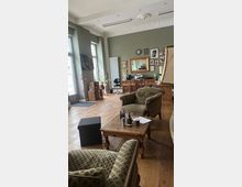 Wohnraum mit Retro-Einrichtung: Ein Raum mit Holzfußboden, grünen Wänden und hoher Decke mit Stuckverzierungen. Zu sehen sind Polstersessel, ein Holztisch mit Deko, ein Schreibtisch, Holzregale und Bilder an der Wand. Große Fenster sorgen für Tageslicht.