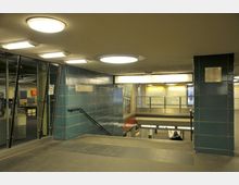 Innenansicht einer U-Bahn-Station mit blauen Fliesenwänden, Treppengeländern und modernen Deckenleuchten. Im Hintergrund befinden sich Bahnsteige, ein Informationsschild und beleuchtete Werbetafeln.