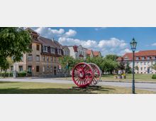 Städtischer Platz mit einer Skulptur aus rot lackierten Eisenbahnrädern in der Mitte, umgeben von historischen Wohngebäuden mit Ziegeldächern. Der Platz ist von Bäumen, Grünflächen und einer klassischen Straßenlaterne geprägt.