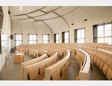 Hörsaal mit halbkreisförmig angeordneten Sitzreihen aus hellem Holz, einem Rednerpult vorne links und großen Fenstern an der Rückseite, die den Raum mit Tageslicht durchfluten. Die Decke ist gewölbt und mit integrierter Beleuchtung versehen.