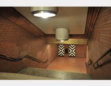 Treppenabgang in einer U-Bahn-Station mit überwiegend roten Backsteinwänden, schwarzen Handläufen und zwei Türen mit markanten schwarz-weißen Zickzackmustern am Ende. Die Szene ist beleuchtet durch runde, große Deckenlampen.