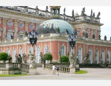 Barocke Palastfassade mit einer großen grünen Kuppel, verziert mit Skulpturen und aufwendigen Verzierungen. Im Vordergrund stehen dekorative Laternen sowie Statuen und symmetrisch platzierte Sträucher, während zwei Personen auf einem gepflasterten Weg spazieren.