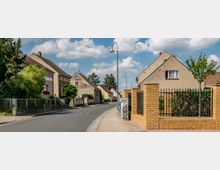 Straßenszene in einem Wohngebiet mit Einfamilienhäusern, teils mit Putzfassaden und Satteldächern. Rechts ist ein Zaun aus gelben Ziegeln und Metallstäben zu sehen, während die Straße und Gehwege mit Laternen gesäumt sind.