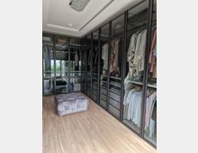 Ein begehbarer Kleiderschrank mit Schränken aus Glasfronten, in denen Kleidung auf Kleiderbügeln und in Schubladen geordnet ist. Der Raum hat einen hellen Holzfußboden, einen gepolsterten Hocker in der Mitte und eine moderne Deckenleuchte.