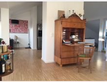 Wohnzimmer mit hellem Holzfußboden, einem antiken Sekretär aus Holz, einem passenden Holzstuhl mit grünem Sitzpolster und moderner Deko. Im Hintergrund sind eine farbige Wanddekoration, ein Stehlampe, ein Sideboard und ein Kühlschrank zu sehen.