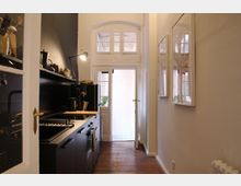 Schmale Küche mit schwarzen Schränken, Spüle und Herd auf der linken Seite. Rechts hängen gerahmte Bilder, im Hintergrund führt eine Glastür mit darüberliegenden Fenstern zu einem hellen Außenbereich.