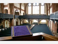 Innenraum einer historischen Kirche mit Backsteinbögen, großen Spitzbogenfenstern und dunklen Holzemporen. Im Vordergrund liegt ein violettes Tuch auf einer Kanzel, die Reihen mit Holzbänken erstrecken sich entlang des Kirchenschiffs.