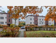 Straßenszene mit Mehrfamilienhäusern im Stil der 1920er Jahre, gekennzeichnet durch helle Fassaden und vertikale Ziegelstreifen. Im Vordergrund Herbstbäume mit orangefarbenem Laub sowie eine Hecke und ein Rasenstück.